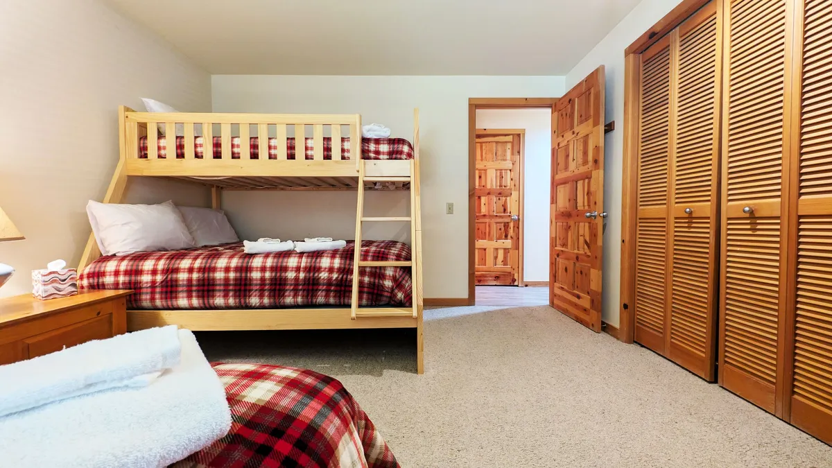 A cheerful bunk room with sturdy wooden bunk beds dressed in plaid bedding, complemented by matching wooden furniture and closet doors. The space feels bright and welcoming, ideal for children or friends sharing a room.