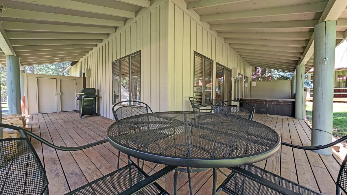 This image features a wooden wrap-around deck furnished with a metal table and black chairs, providing ample seating for outdoor dining. A hot tub is visible to one side, and large windows connect the deck to the interior. The space is well-suited to gatherings and casual relaxation, with room for grilling and enjoying the outdoors.
