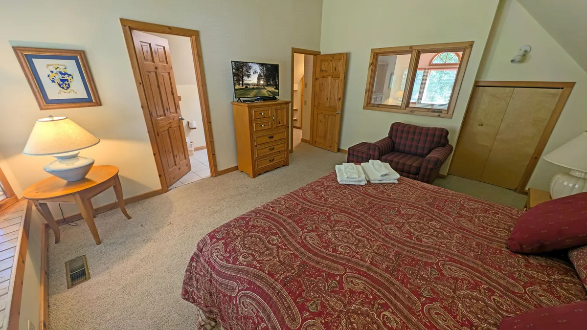 A large, inviting bedroom featuring a king-sized bed with a patterned bedspread, a comfortable armchair by the window, a wooden dresser topped with a TV, and framed artwork on the wall. The room feels cozy and private, perfect for relaxation after a long day.