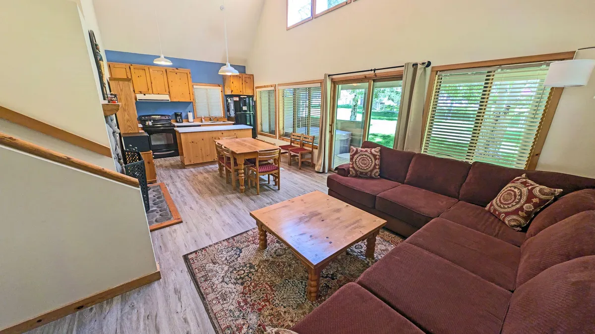 This image showcases the open-concept living space with a welcoming sofa, a dining table with seating, a visible kitchen area, and large windows that flood the space with natural light. The layout encourages gathering, relaxation, and seamless flow between living, dining, and kitchen spaces.