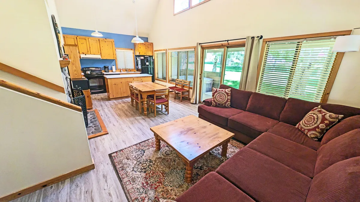 The image showcases a warm, open-concept living space featuring a comfortable sofa, a dining table set for meals, and a modern kitchen—all bathed in natural light from large windows. The scene conveys a spacious, inviting atmosphere ideal for families or groups to relax, gather, and connect.
