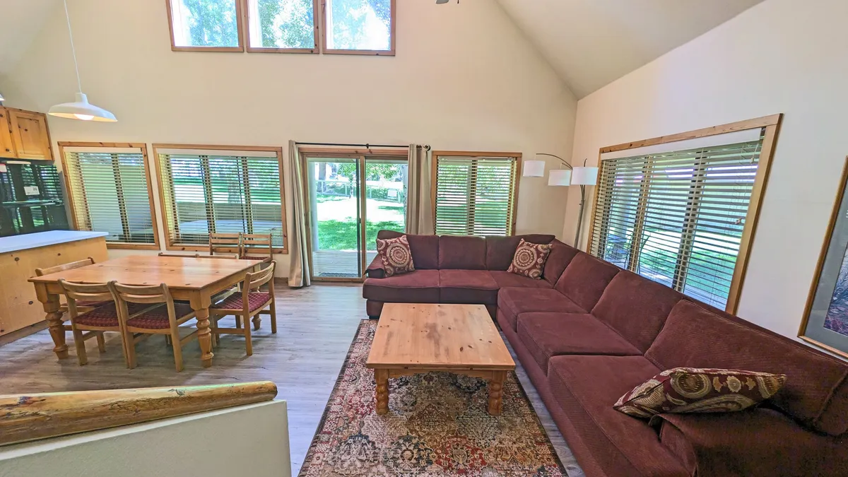 This image features a large sectional sofa arranged around a wooden coffee table and is adjacent to a dining area. The layout suggests a spacious, open-plan living space ideal for group gatherings, casual dining, and shared activities such as playing games or hosting holiday meals. Large windows in the background hint at a connection to the outdoors, further supporting group socialization and relaxation.