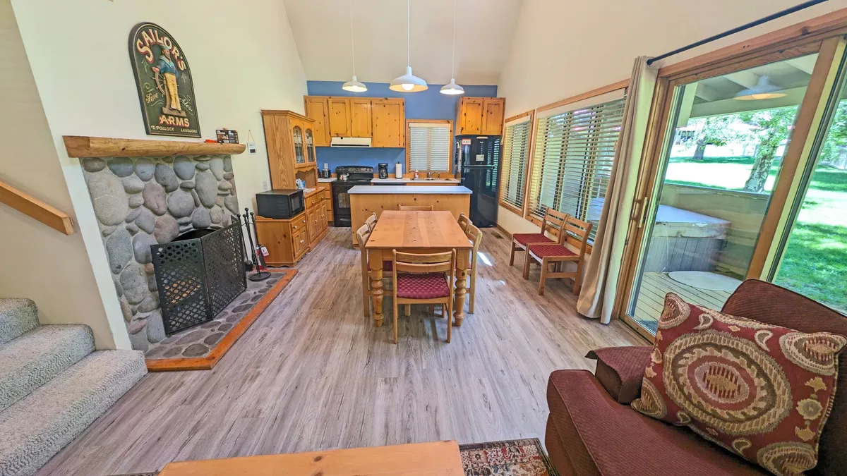 The image centers on a cozy scene featuring a couch, dining table, kitchen island, and a prominent rock-faced fireplace. The setup suggests warmth and togetherness, ideal for family gatherings and cozy evenings by the fire.