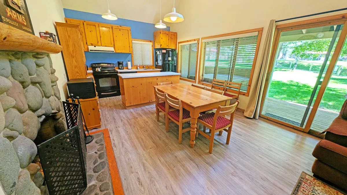 The scene captures a dining table set near a kitchen counter and a prominent stone fireplace, with large windows allowing natural light to fill the room. The setup is inviting for shared meals, group conversations, and entertaining. The combination of dining and living amenities underscores the condo's suitability for memorable group experiences.